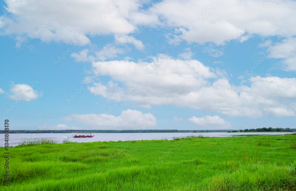 A scenic view of a long tail boat driver and tourists sailing in river with green lawn grass filed and trees against blue sky with cloud, presenting a breathtaking scenery in Bueng Kan, Thailand.