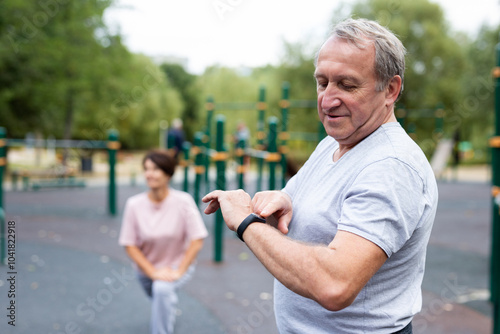 Ssenior man in sportswear looks at fitness watch or at pedometer while exercising in city park photo