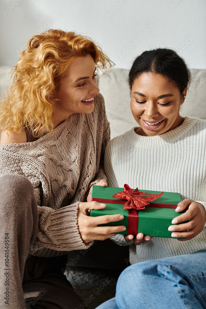 Two women express happiness while exchanging a wrapped gift in their cozy living space. Two women express happiness while exchanging a wrapped gift in their cozy living space.