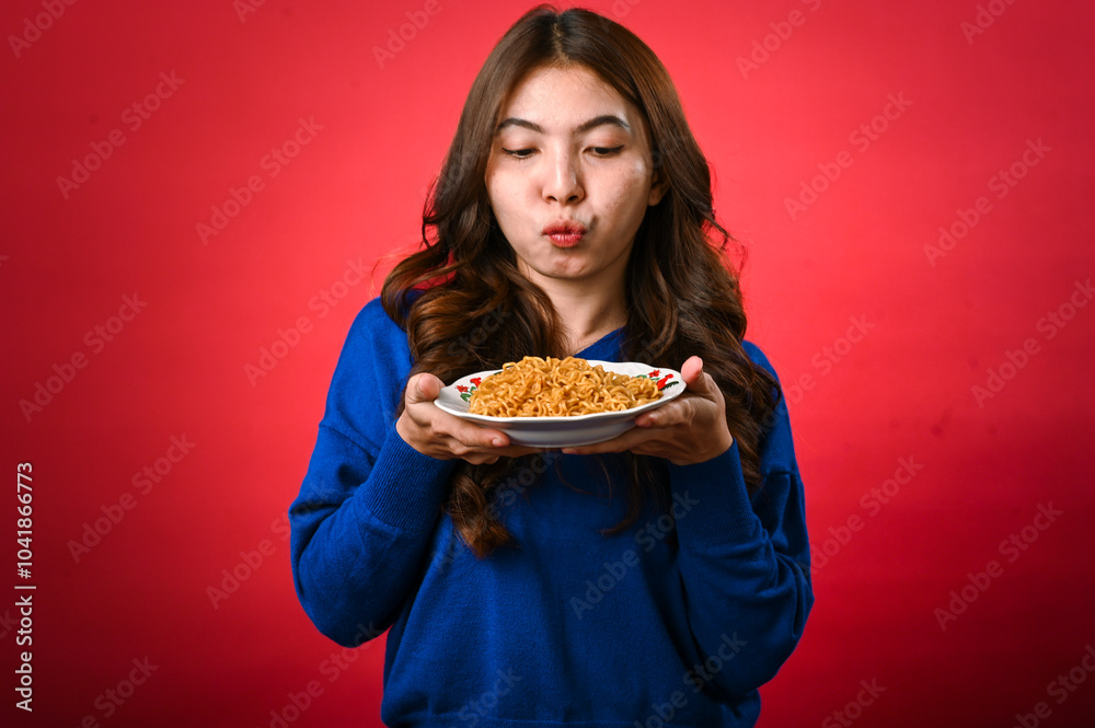 An Asian woman in a blue sweater holds a plate of noodles close to her face, licking her lips in anticipation. She stands against a red background with a playful and hungry expression An Asian woman in a blue sweater holds a plate of noodles close to her face, licking her lips in anticipation. She stands against a red background with a playful and hungry expression
