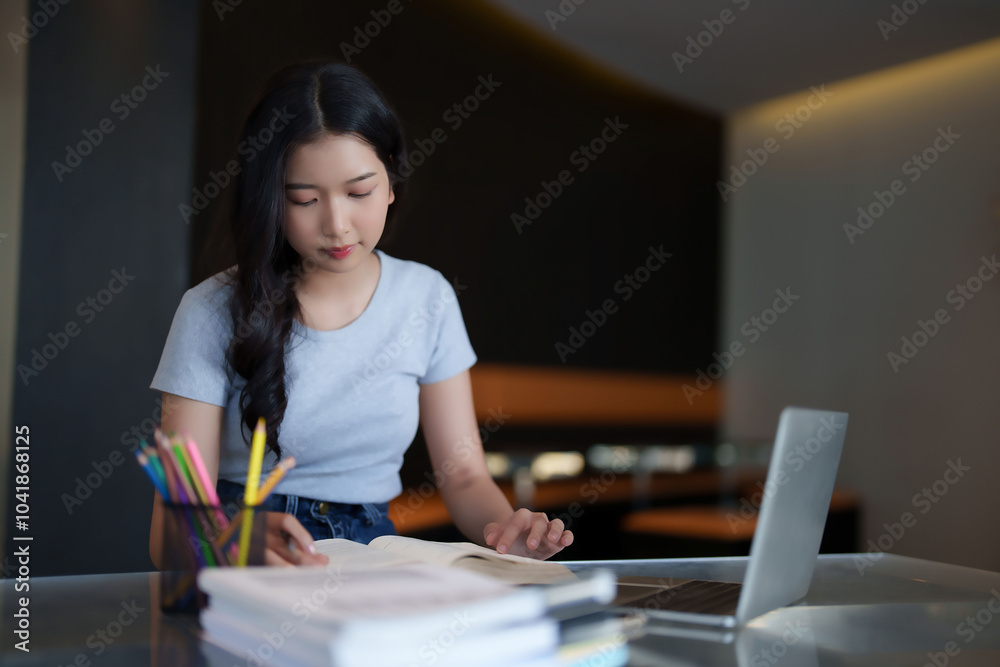 Asian teenage student woman reading a book and writing notes to doing homework while watching lecture video online lesson on digital laptop to studying and learning about education knowledge in cafe Asian teenage student woman reading a book and writing notes to doing homework while watching lecture video online lesson on digital laptop to studying and learning about education knowledge in cafe