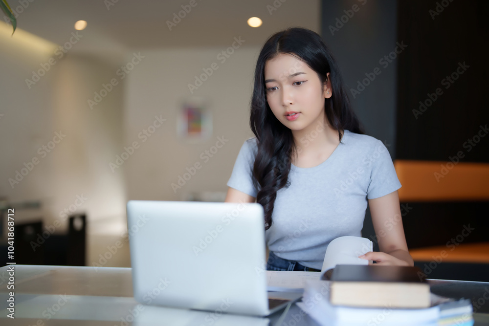 Asian teenage student woman reading a book and typing to researching data on laptop for doing assignment homework after watching lecture video online lesson and studying education knowledge in cafe Asian teenage student woman reading a book and typing to researching data on laptop for doing assignment homework after watching lecture video online lesson and studying education knowledge in cafe
