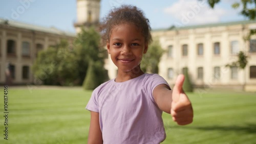 Portrait happy smiling African American little student pupil girl daughter schoolgirl female child kid city outdoor studying elementary school education college academy showing thumb up good recommend photo