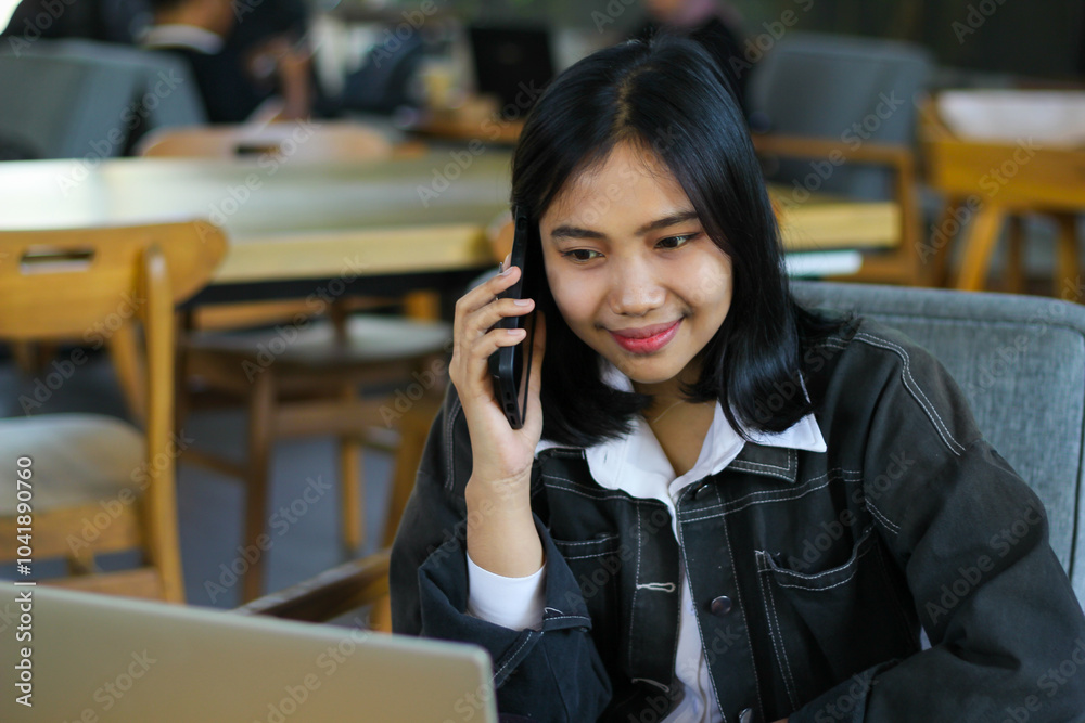 happy asian woman make mobile phone call while working in indoor cafe with laptop, looking at laptop screen happy asian woman make mobile phone call while working in indoor cafe with laptop, looking at laptop screen