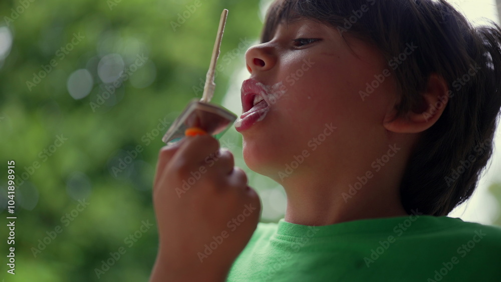 Young boy enjoying ice cream, licking the popsicle in an upward motion. Close-up of a child savoring the dessert outdoors against a green natural background on a warm day Young boy enjoying ice cream, licking the popsicle in an upward motion. Close-up of a child savoring the dessert outdoors against a green natural background on a warm day