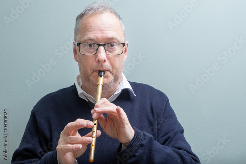 Experienced senior man, with greying hair, casually dressed, wearing dark glasses, playing a gold or brass coloured tin whistle, penny whistle, traditional Irish musical instrument.