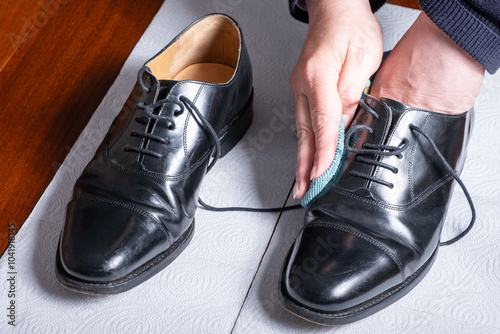 Close up view of man cleaning black brogue, hard leather shoes with microfibre cloth on wooden table, with white kitchen roll protective paper below the shoes.