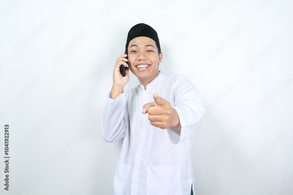 cheerful muslim asian man pointing forward to camera while speaking on his phone with smiling face isolated cheerful muslim asian man pointing forward to camera while speaking on his phone with smiling face isolated