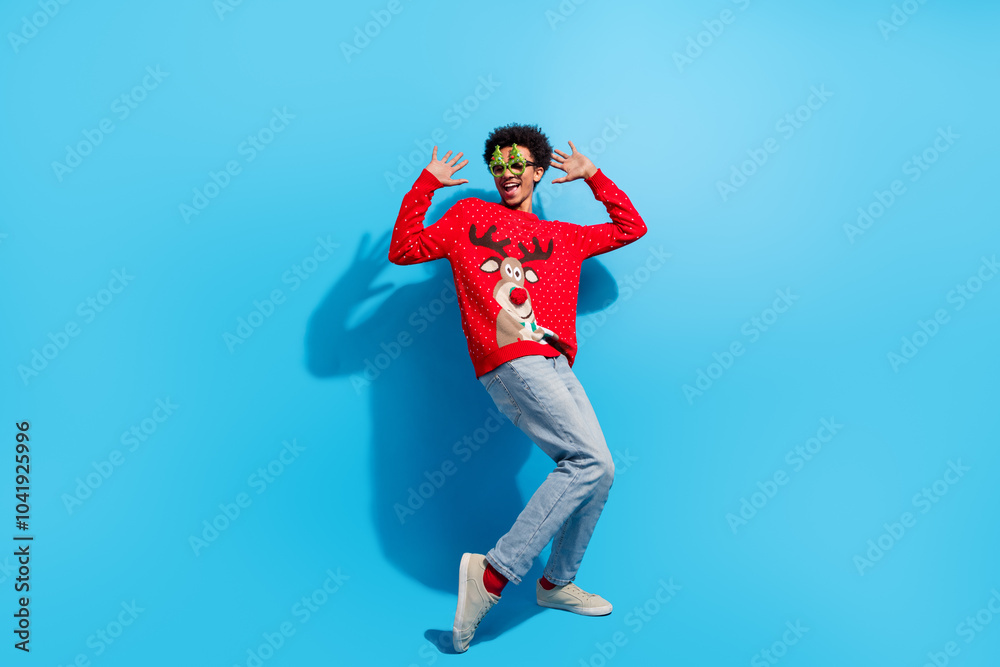 Full body photo of attractive young man excited dancing dressed red christmas sweater theme party on blue color background Full body photo of attractive young man excited dancing dressed red christmas sweater theme party on blue color background