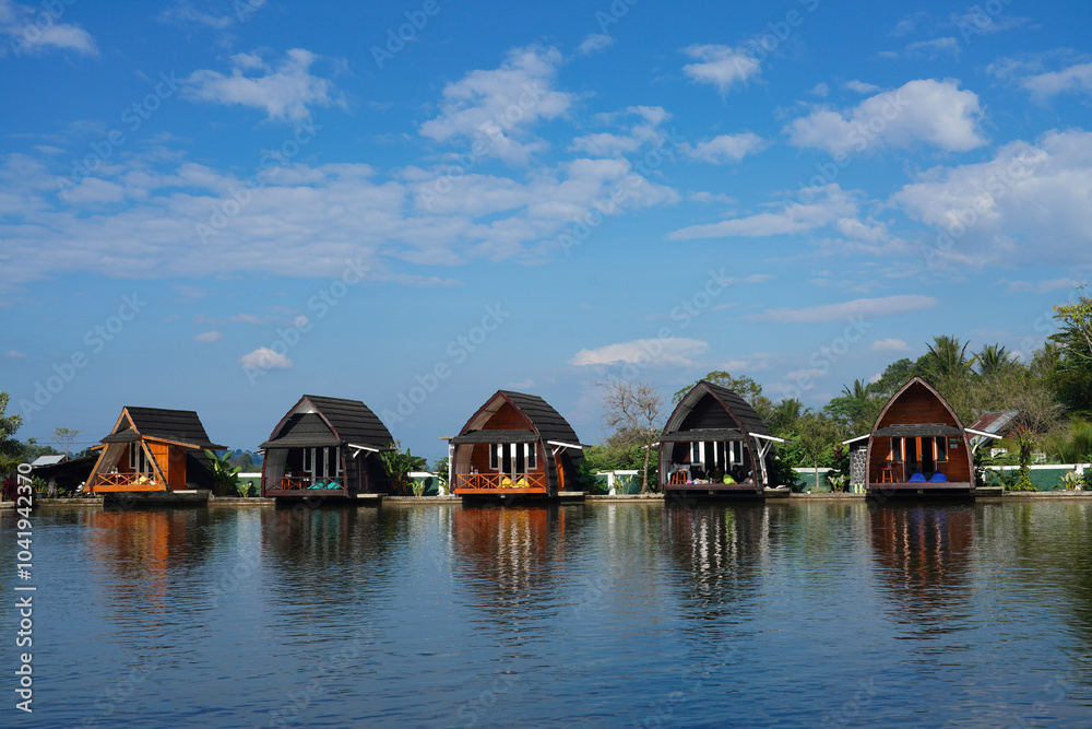 Naklejka premium Traditional wooden cottages line the lake shore, with the reflection of the calm water. Bright blue sky. Beautiful view of the lakeside villas