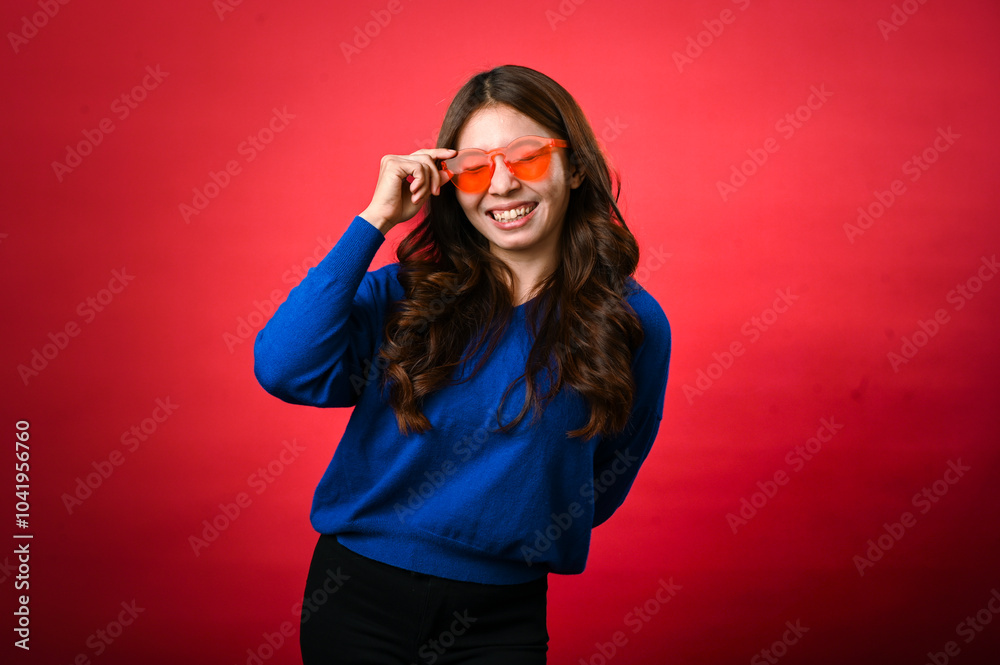 An Asian woman wearing a blue sweater is smiling while adjusting her bright orange sunglasses. She is standing against a red background, giving a playful and confident expression An Asian woman wearing a blue sweater is smiling while adjusting her bright orange sunglasses. She is standing against a red background, giving a playful and confident expression