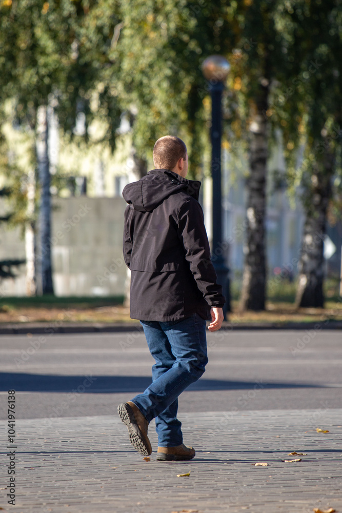 A man wearing a black jacket and blue jeans walks down a sidewalk A man wearing a black jacket and blue jeans walks down a sidewalk