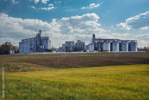 large grain elevator with multiple cylindrical metal silos, structural elements, and a complex system of pipes. It stands against a blue sky, with a field in the foreground after harvest