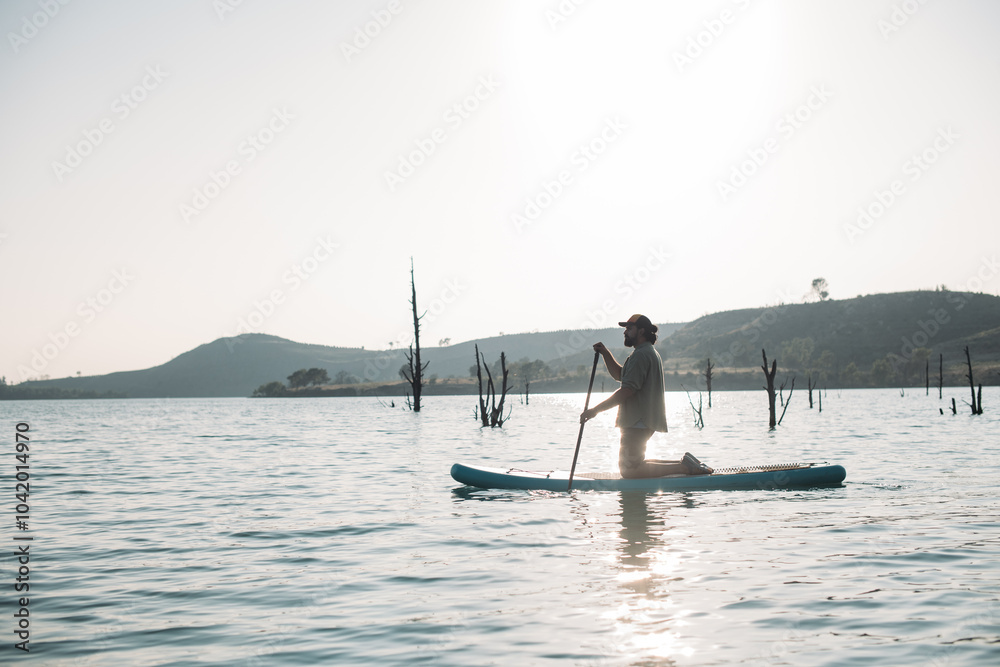 Naklejka premium A man is surfing on a sap board on calm water at sunset.