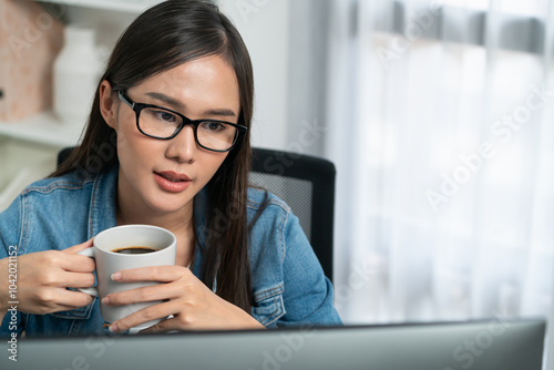 Portrait working young asian creative manager with glasses focusing on pc while drinking hot coffee, creating with website design on social media with blue jeans shirt at home office. Stratagem.