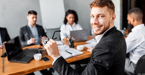 Young Entrepreneur Smiling Looking At Camera Posing Sitting During Business Partners Meeting In Modern Office.