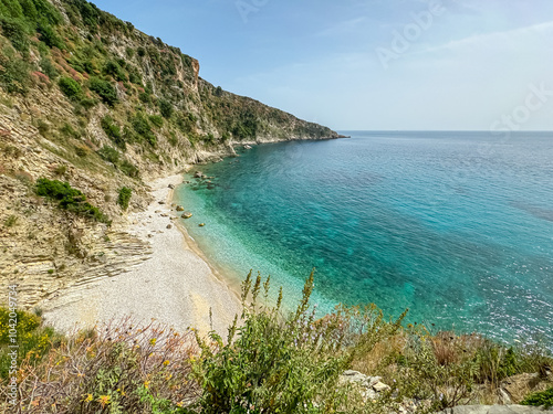 Selective focus on flowers with scenic view of secluded pebble beach Filikuri surrounded by dramatic cliffs in Himare, Albania. Crystal-clear turquoise water of Ionian Mediterranean sea. Tranquility photo