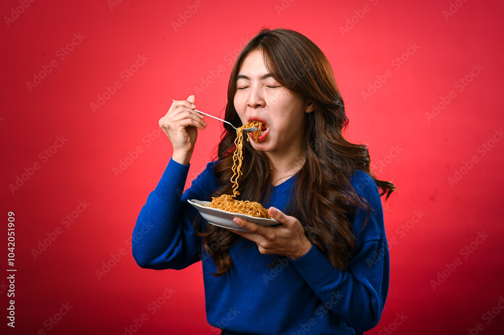An Asian woman in a blue sweater is enjoying a plate of noodles, lifting a forkful to her mouth. She stands against a bold red background, fully focused on her delicious meal. An Asian woman in a blue sweater is enjoying a plate of noodles, lifting a forkful to her mouth. She stands against a bold red background, fully focused on her delicious meal.