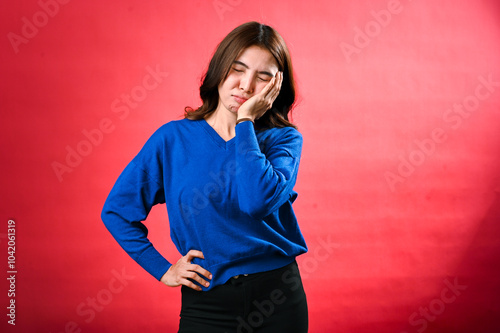 An Asian woman with long brown hair, wearing a blue sweater, is wincing in pain with her hand on her cheek and her other hand on her hip. She stands against a red background, looking frustrated