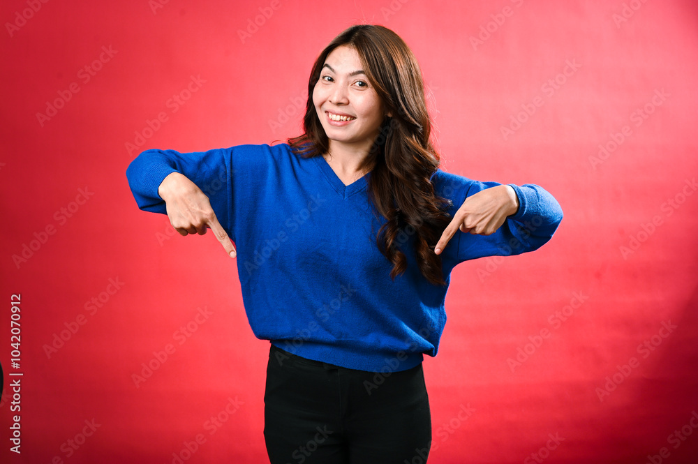 An Asian woman with long brown hair smiles while pointing downwards with both hands. She wears a blue sweater, posing against a bold red background, exuding a confident and friendly vibe An Asian woman with long brown hair smiles while pointing downwards with both hands. She wears a blue sweater, posing against a bold red background, exuding a confident and friendly vibe