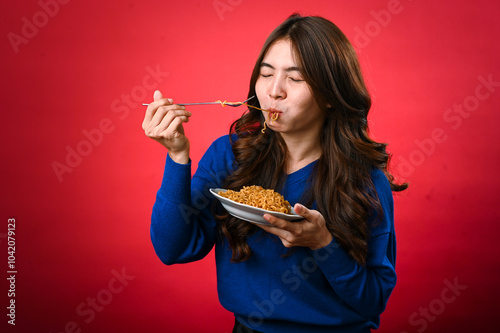 An Asian woman in a blue sweater is enjoying a plate of noodles, lifting a forkful to her mouth. She stands against a bold red background, fully focused on her delicious meal. photo