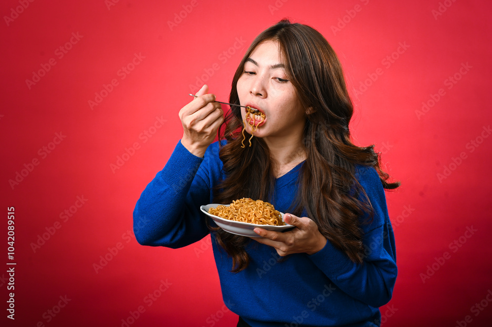 An Asian woman in a blue sweater is enjoying a plate of noodles, lifting a forkful to her mouth. She stands against a bold red background, fully focused on her delicious meal. An Asian woman in a blue sweater is enjoying a plate of noodles, lifting a forkful to her mouth. She stands against a bold red background, fully focused on her delicious meal.