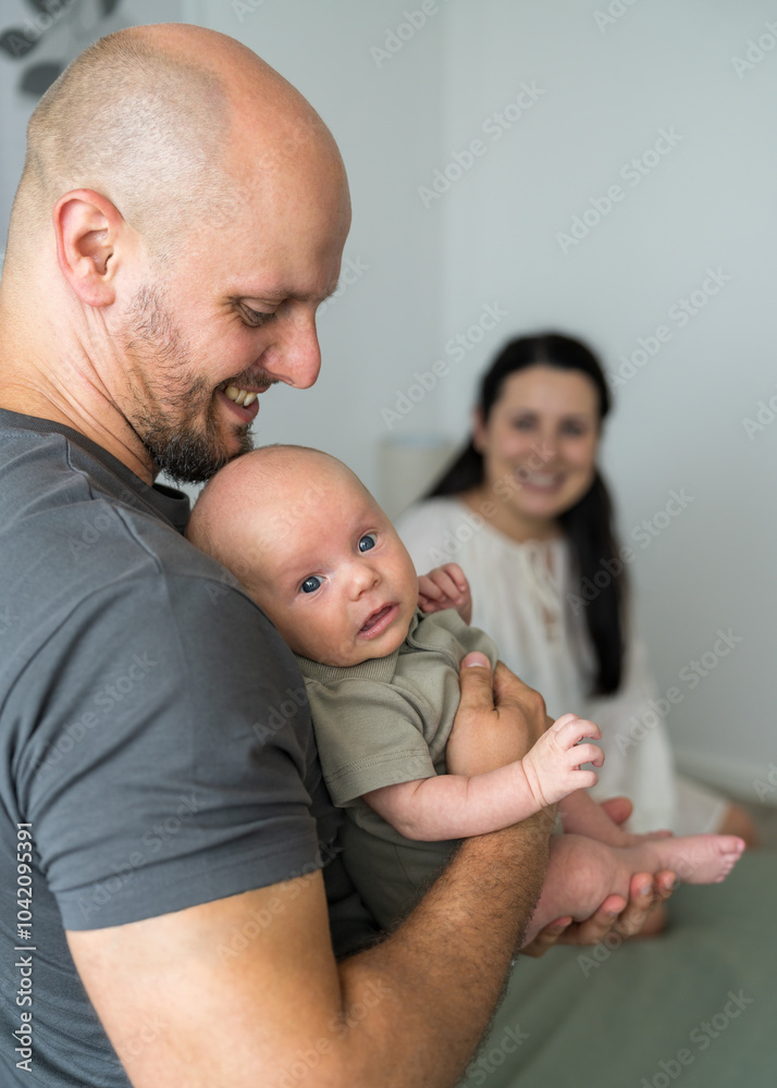 In foreground is smiling dad with newborn baby in arms. Mom sits on bed and looks at loved ones. In center of attention is man. Happy family. In foreground is smiling dad with newborn baby in arms. Mom sits on bed and looks at loved ones. In center of attention is man. Happy family.
