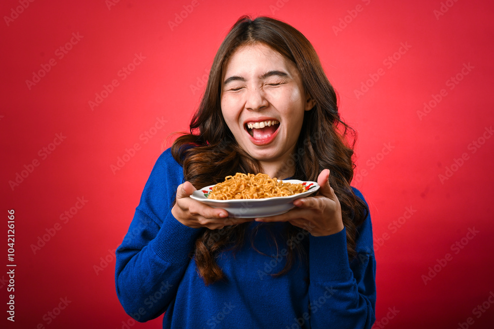 An Asian woman in a blue sweater holds a plate of noodles close to her face, licking her lips in anticipation. She stands against a red background with a playful and hungry expression An Asian woman in a blue sweater holds a plate of noodles close to her face, licking her lips in anticipation. She stands against a red background with a playful and hungry expression
