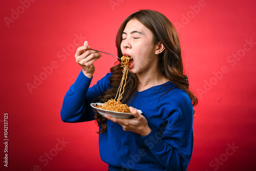 An Asian woman in a blue sweater is enjoying a plate of noodles, lifting a forkful to her mouth. She stands against a bold red background, fully focused on her delicious meal.