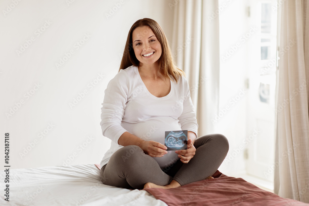 A pregnant woman smiles brightly while sitting on a bed, holding an ultrasound image. The soft light from the window creates a warm and inviting atmosphere in the room. A pregnant woman smiles brightly while sitting on a bed, holding an ultrasound image. The soft light from the window creates a warm and inviting atmosphere in the room.