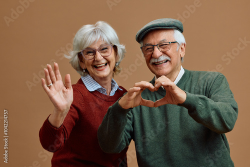 Happy senior man making heart shape with hands while his wife is waving to camera.