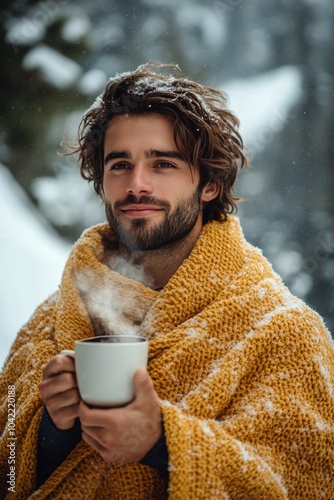 Man wrapped in a yellow blanket enjoys a warm drink in a snowy forest during ...