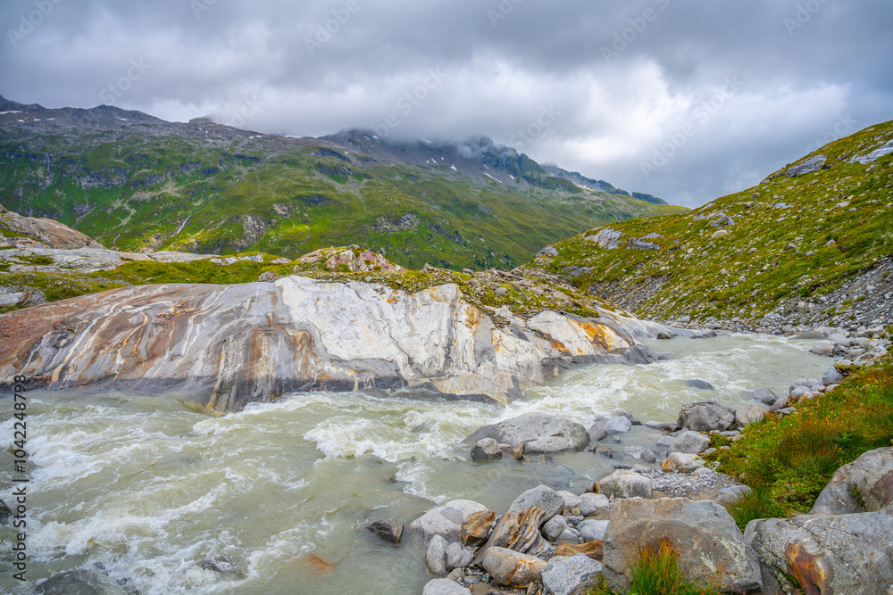 The Schlaten Glacier stream winds through the rocky landscape of Hohe Tauern National Park, surrounded by lush greenery and majestic mountains under a cloudy sky. The Schlaten Glacier stream winds through the rocky landscape of Hohe Tauern National Park, surrounded by lush greenery and majestic mountains under a cloudy sky.