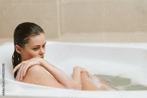 Thoughtful young woman relaxing in a bathtub surrounded by bubbles, enjoying a moment of solitude and self-care in a serene bathroom setting.