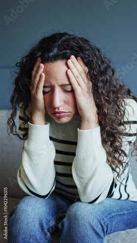 Young woman sitting on a sofa at home, suffering from a headache and displaying signs of stress and discomfort. Woman suffering from headache sitting on couch