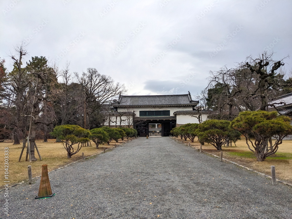 Kyoto, Japan - 2023.12.16: Nijo Castle Kita Otemon in Nijo Castle with a treelined footpath leading to the brick and wooden structure with traditional roof tiles under a cloudy sky Kyoto, Japan - 2023.12.16: Nijo Castle Kita Otemon in Nijo Castle with a treelined footpath leading to the brick and wooden structure with traditional roof tiles under a cloudy sky