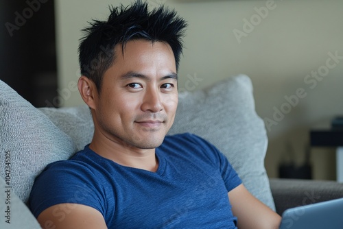 Man with short black hair, wearing a blue shirt, sitting on a couch with a laptop, casual home setting, relaxed expression, side view 2 photo