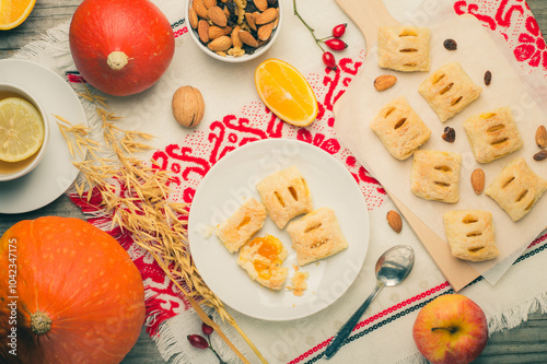 Handcrafted pastries with dried fruits and nuts, paired with fresh produce. Golden puff pastries with orange filling. The wooden background and folk tablecloth. Top view, flat lay. photo