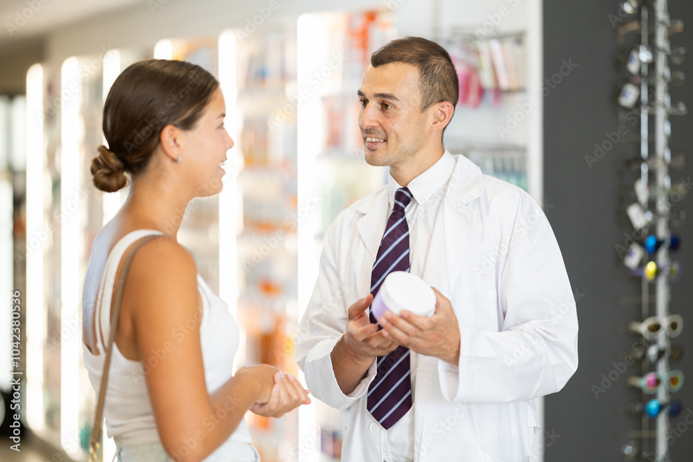 Obraz premium Positive man apothecary speaking to female customer holding cream in hand in pharmacy store