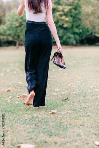 Beautiful graceful gentle happy free french girl with long dark hair and elegant trousers runs barefoot on the grass and holds her high-heeled shoes in her hands photo