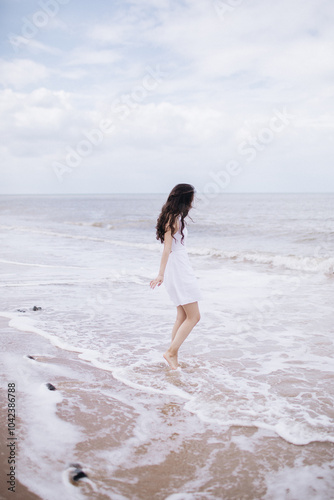 Beautiful graceful gentle girl with long dark hair in a white dress stands on the seashore, the waves wash her bare feet, her hair flutters in the wind