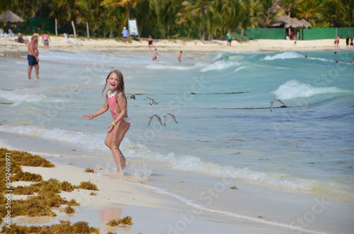 Menina feliz, vestindo traje de banho, correndo na beira do mar, com pássaros ao fundo e o mar azul do caribe com algumas ondas photo