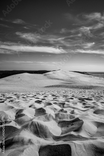Black and white photo of sand dunes of Pilat with contrasty shadows creating patterns. The sky is partly cloudy, and small figures are visible on the dunes