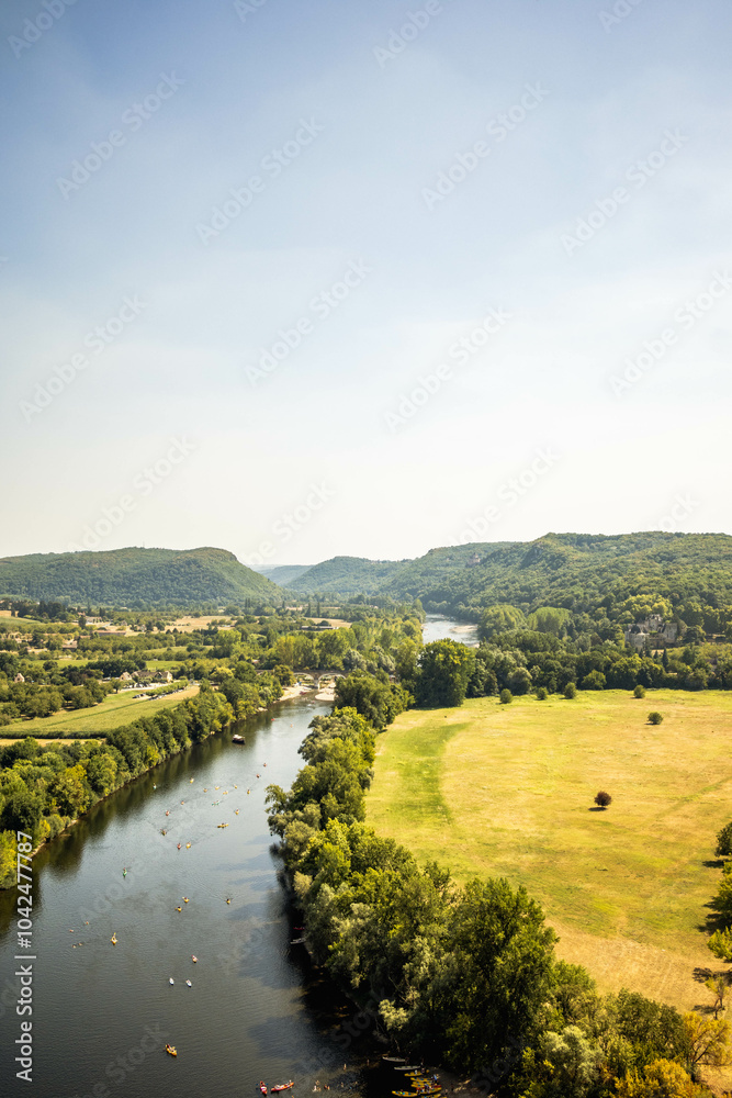 Obraz premium A river winding through a lush green valley, bordered by trees on one side and an open grassy field on the other. Small boats or kayaks are visible on the river under a clear, sunny sky