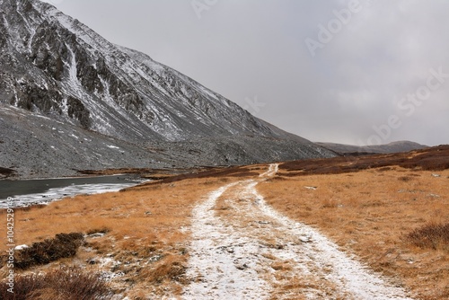 A field road sprinkled with the first snow goes through a clearing lying at the foot of a rocky mountain range on a cloudy autumn day.