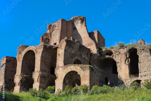 Ruins of the Temple of Apollo Palatine: Ancient Roman temple with crumbling brick walls, arches, and columns, set against a clear blue sky. Lush greenery covers the surrounding hills.