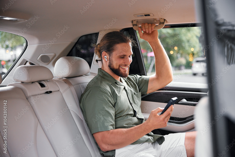 Obraz premium Man using smartphone in the back seat of a car, smiling happily while wearing earbuds