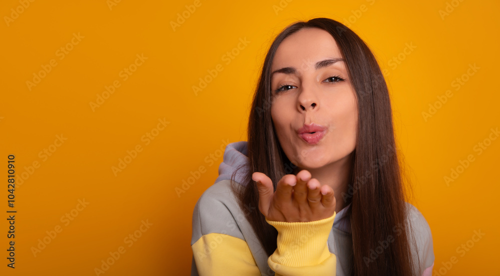 A close-up portrait of a young woman blowing a kiss towards the camera against a bright yellow background. She wears a pastel hoodie, conveying a playful, affectionate, and cheerful vibe. A close-up portrait of a young woman blowing a kiss towards the camera against a bright yellow background. She wears a pastel hoodie, conveying a playful, affectionate, and cheerful vibe.