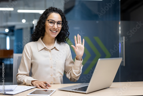 Hispanic woman in office waving at camera, seated at desk with laptop, phone, documents. Reflects friendly work environment, professional interaction, positive communication, confident demeanor.