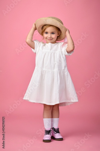 Little cute girl in white summer dress in hat and shoes, knee socks posing in studio on pink background